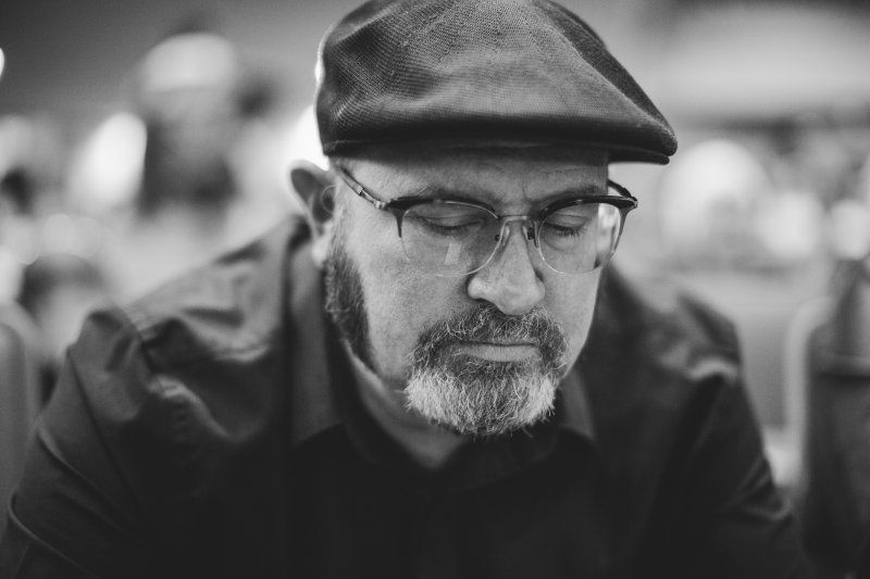 A man with a beard is wearing a flat cap and glasses. He is looking down, in the midst of prayer.